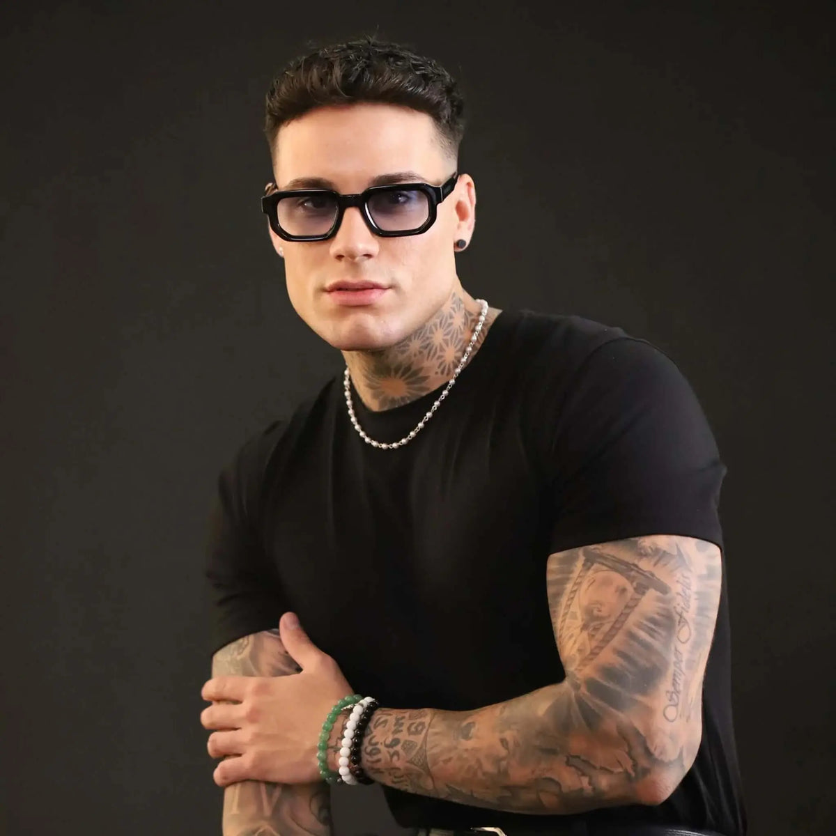 Gafas de sol con montura cuadrada negra de acetato y lentes en tono azul claro sobre un hombre con tatuajes.