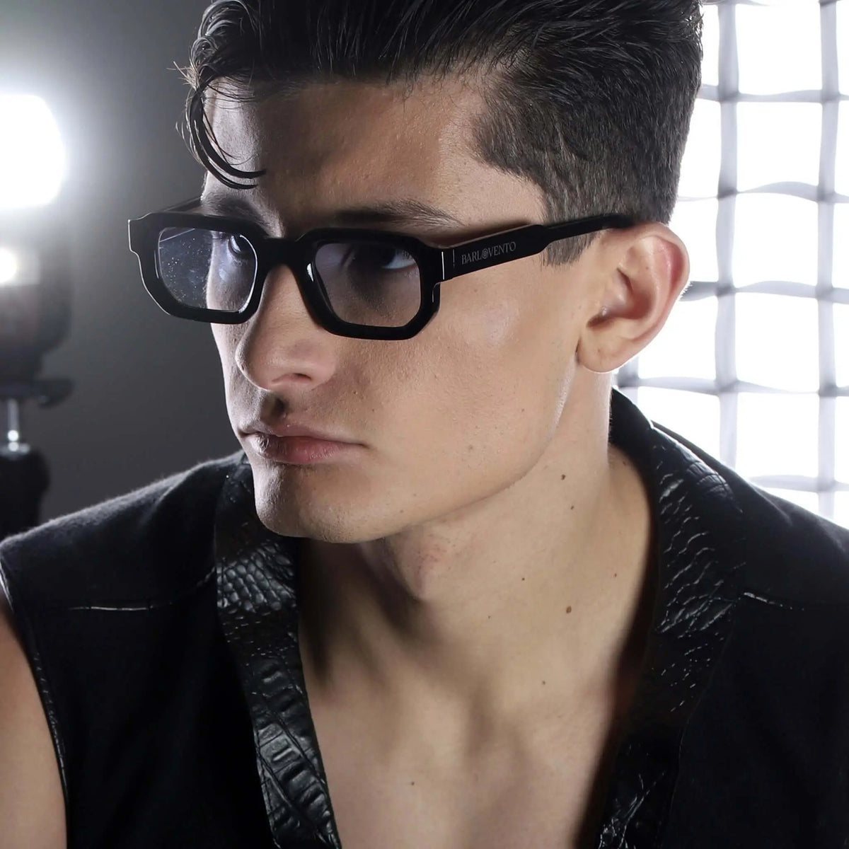Gafas de sol rectangulares con montura negra gruesa y lentes azuladas, usadas por un hombre con chaleco negro texturizado.