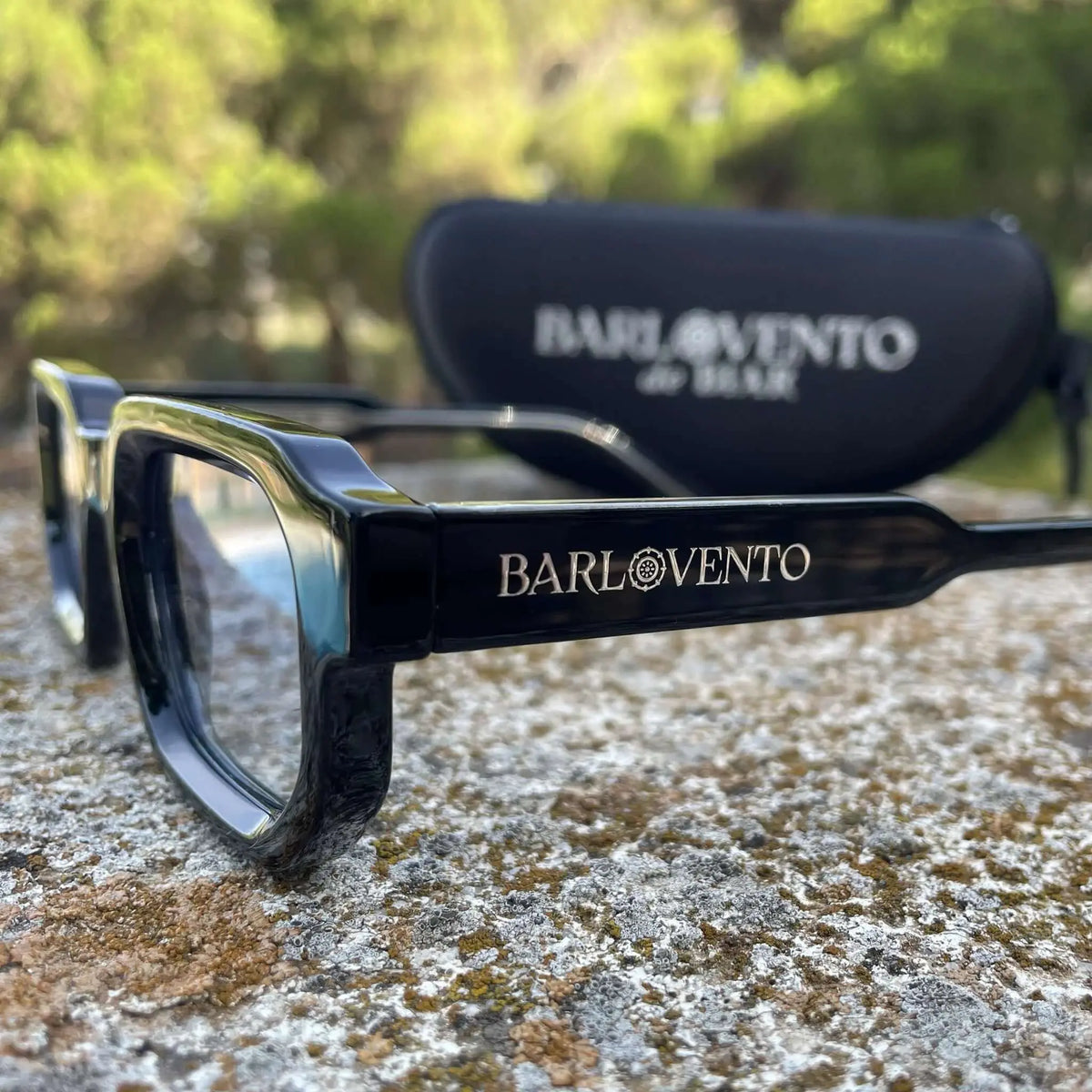 Gafas de sol negras de estilo rectangular con lentes azuladas, apoyadas sobre una superficie de piedra rústica.