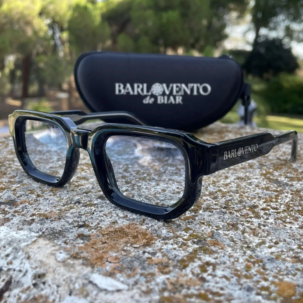 Gafas de sol negras de estilo rectangular con lentes azules claras, sobre superficie de piedra y estuche de fondo.