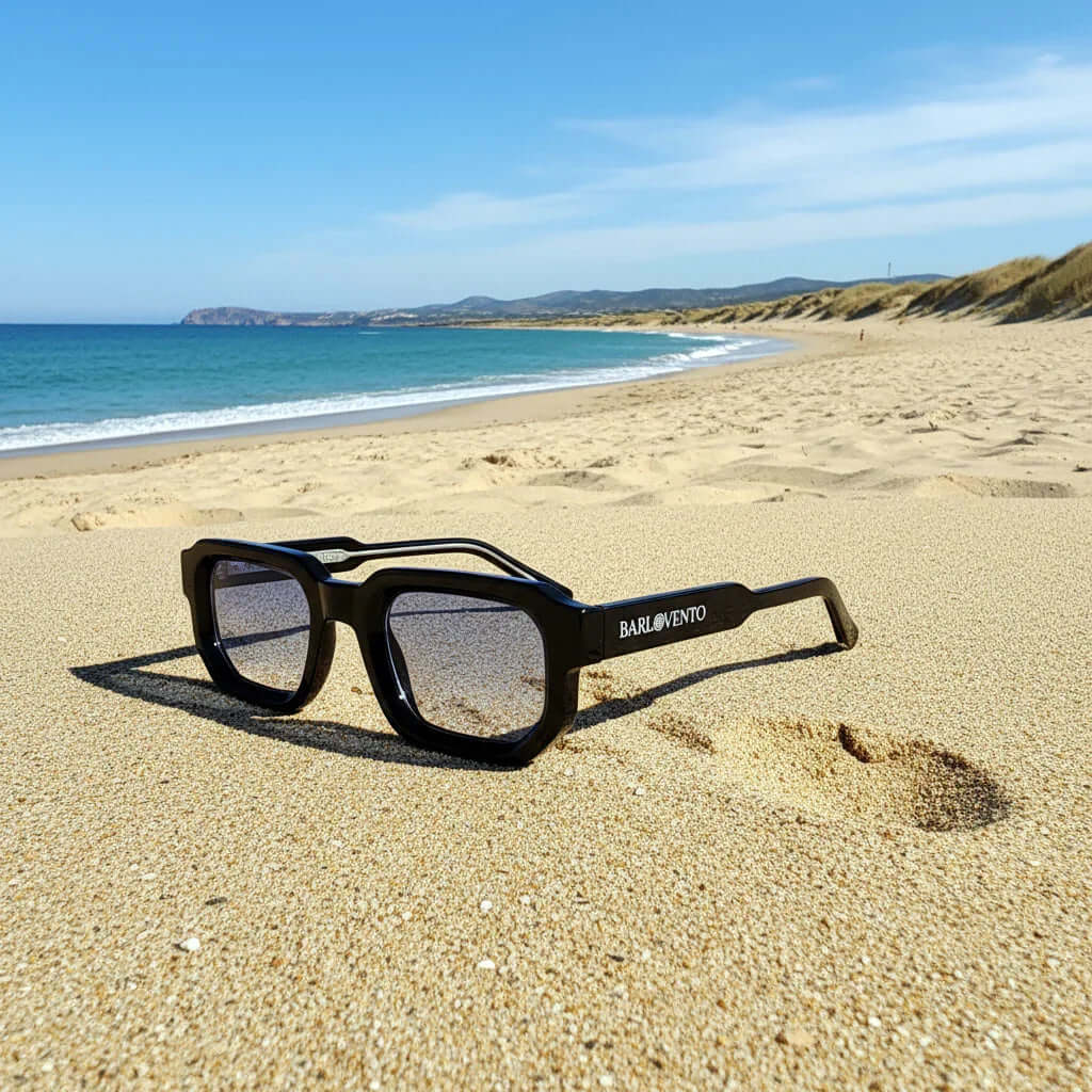 Gafas de sol negras de estilo rectangular con lentes azules sobre la arena de una playa.