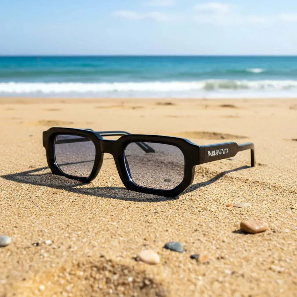 Gafas de sol negras de estilo rectangular con cristales azulados sobre la arena de una playa.