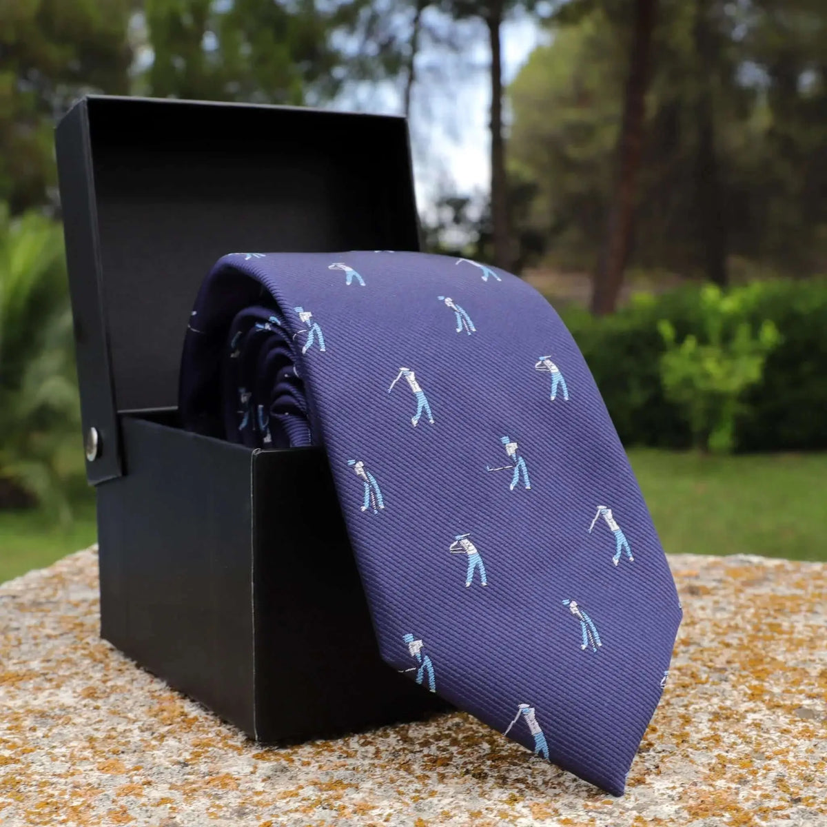 Corbata azul marino de seda con estampado de golfistas en caja de regalo negra sobre superficie de piedra.