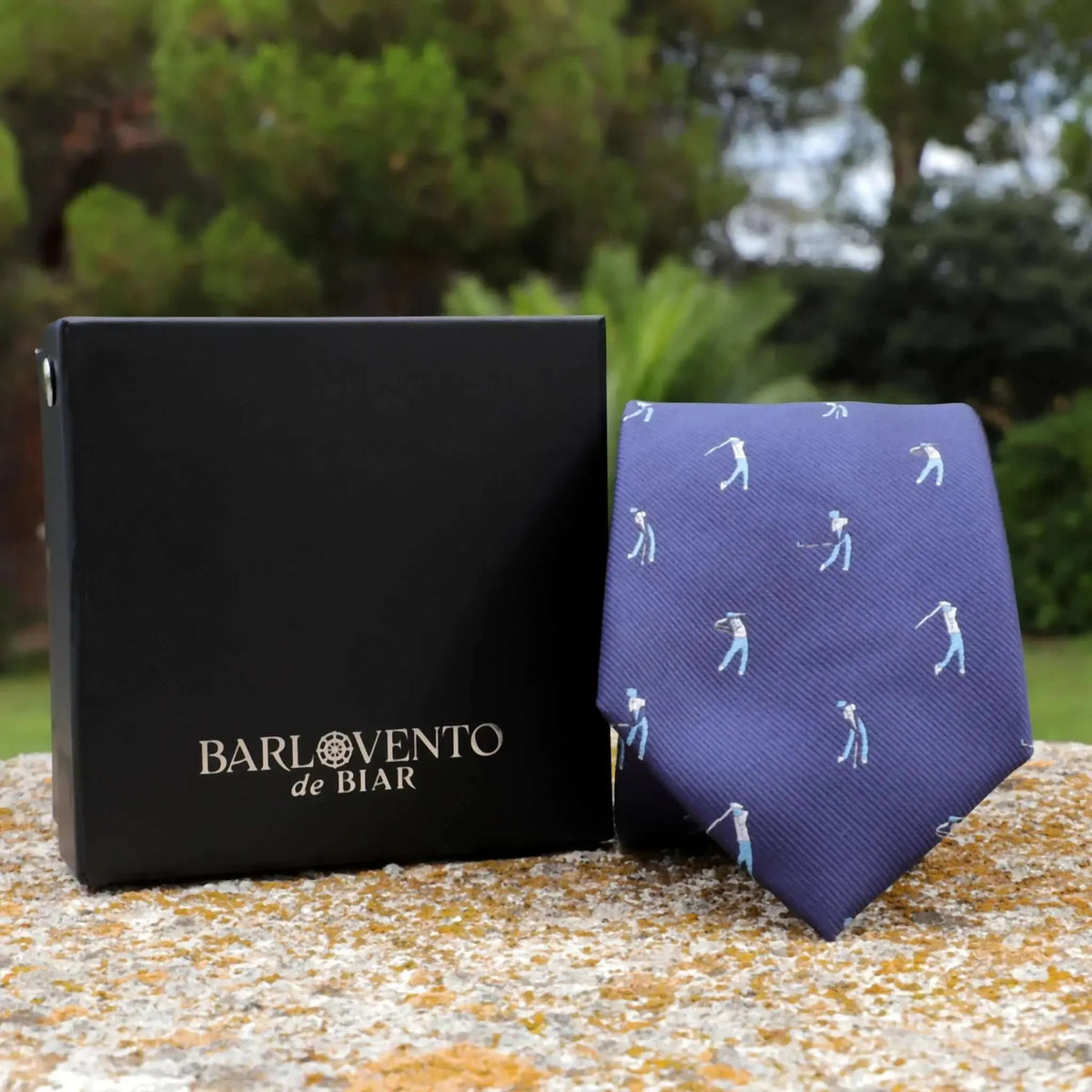 Corbata azul con estampado de golfistas en miniatura y acabado satinado, junto a caja negra de regalo sobre piedra.