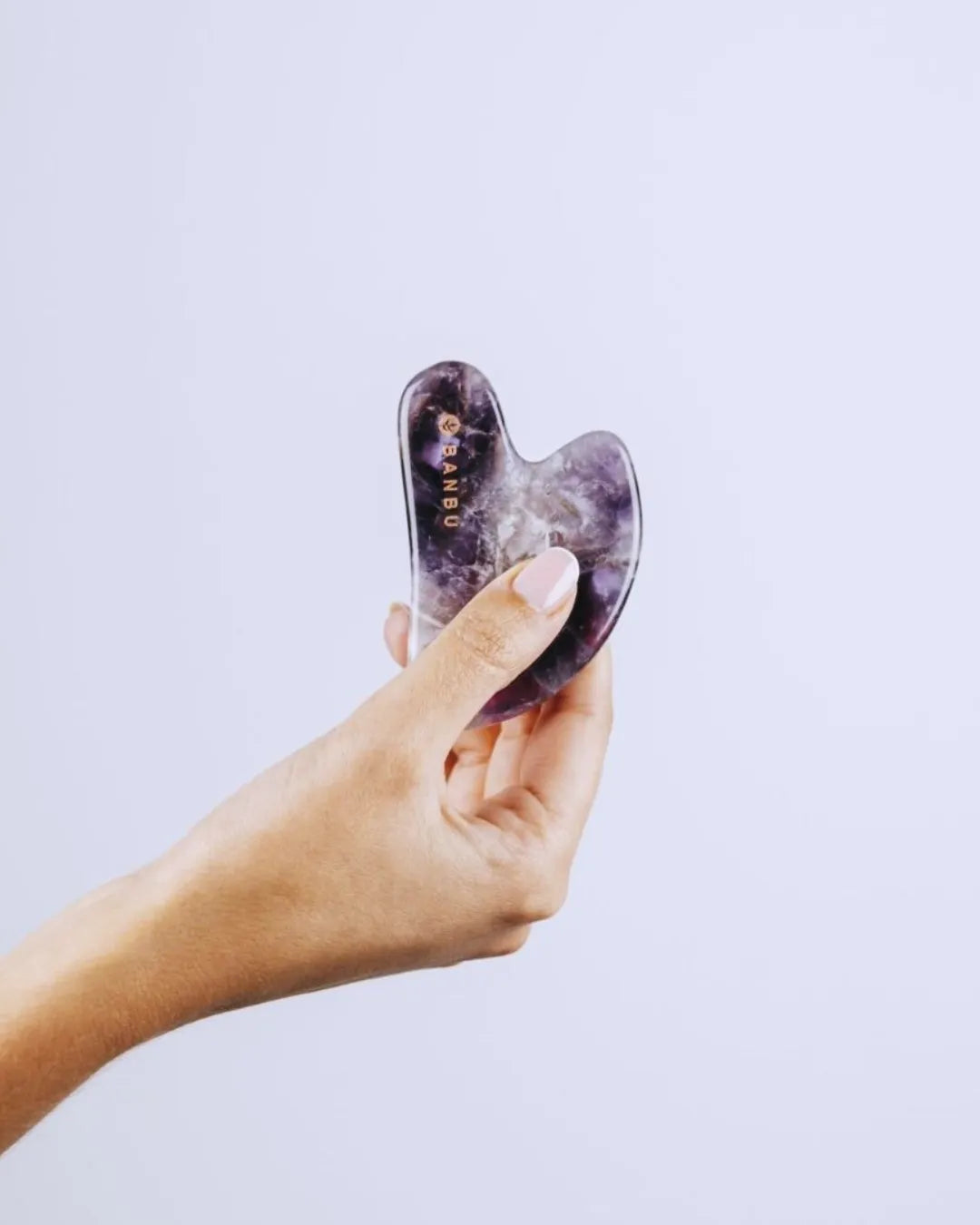 Mano sosteniendo una herramienta Gua Sha de amatista natural con vetas moradas y logo de la marca BANBU grabado.