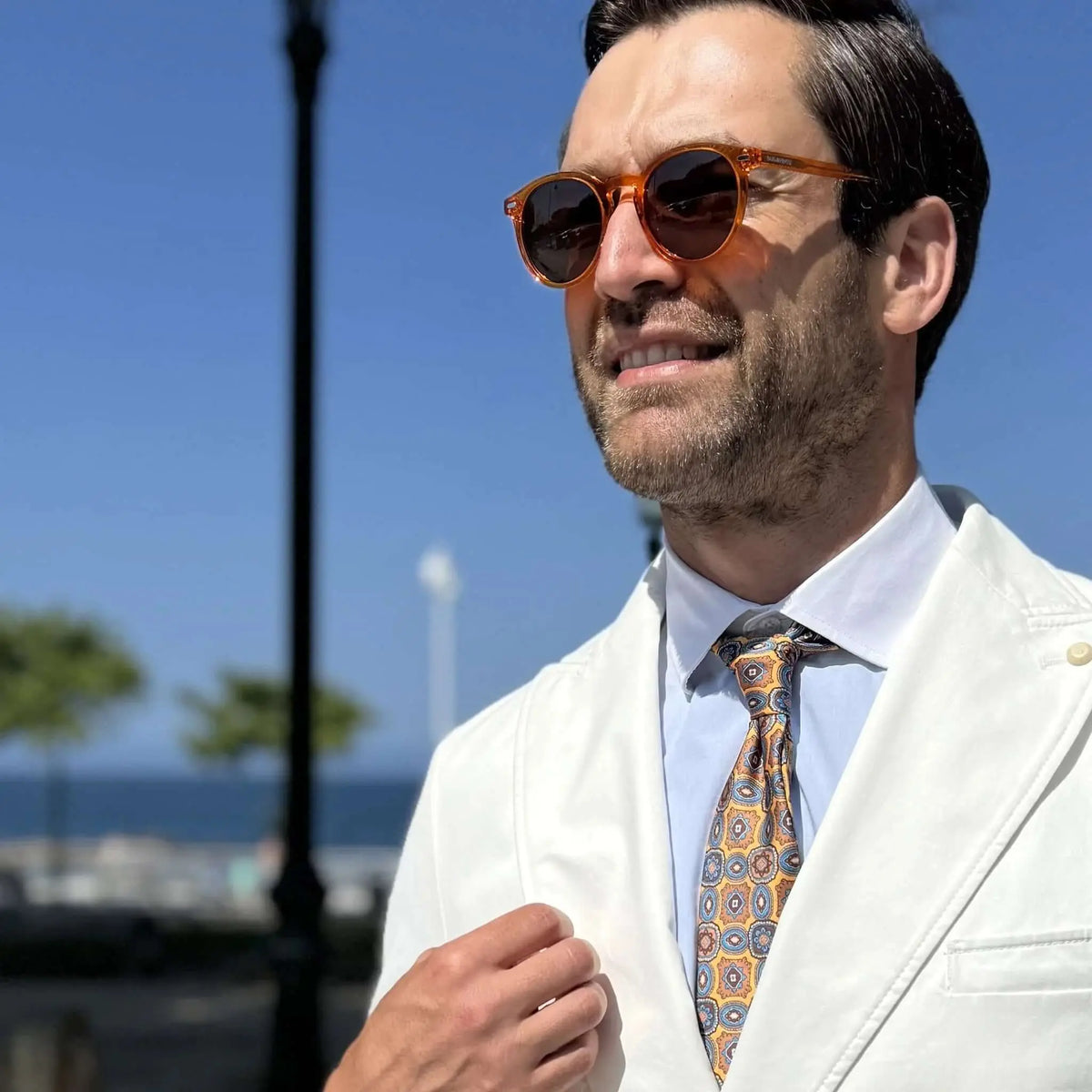 Hombre con traje blanco y corbata usando gafas de sol redondas de acetato color naranja cristal y lentes oscuros.