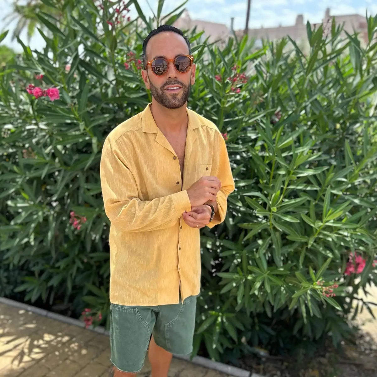 Hombre con camisa amarilla y gafas de sol redondas de montura naranja translúcida frente a un arbusto verde.