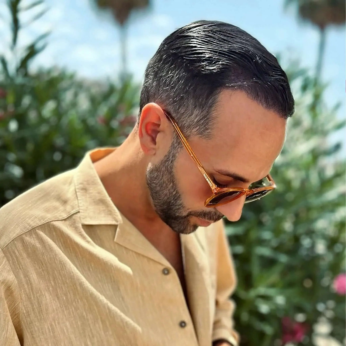 Hombre con camisa beige y gafas de sol de pasta naranja transparente estilo retro en exterior soleado.