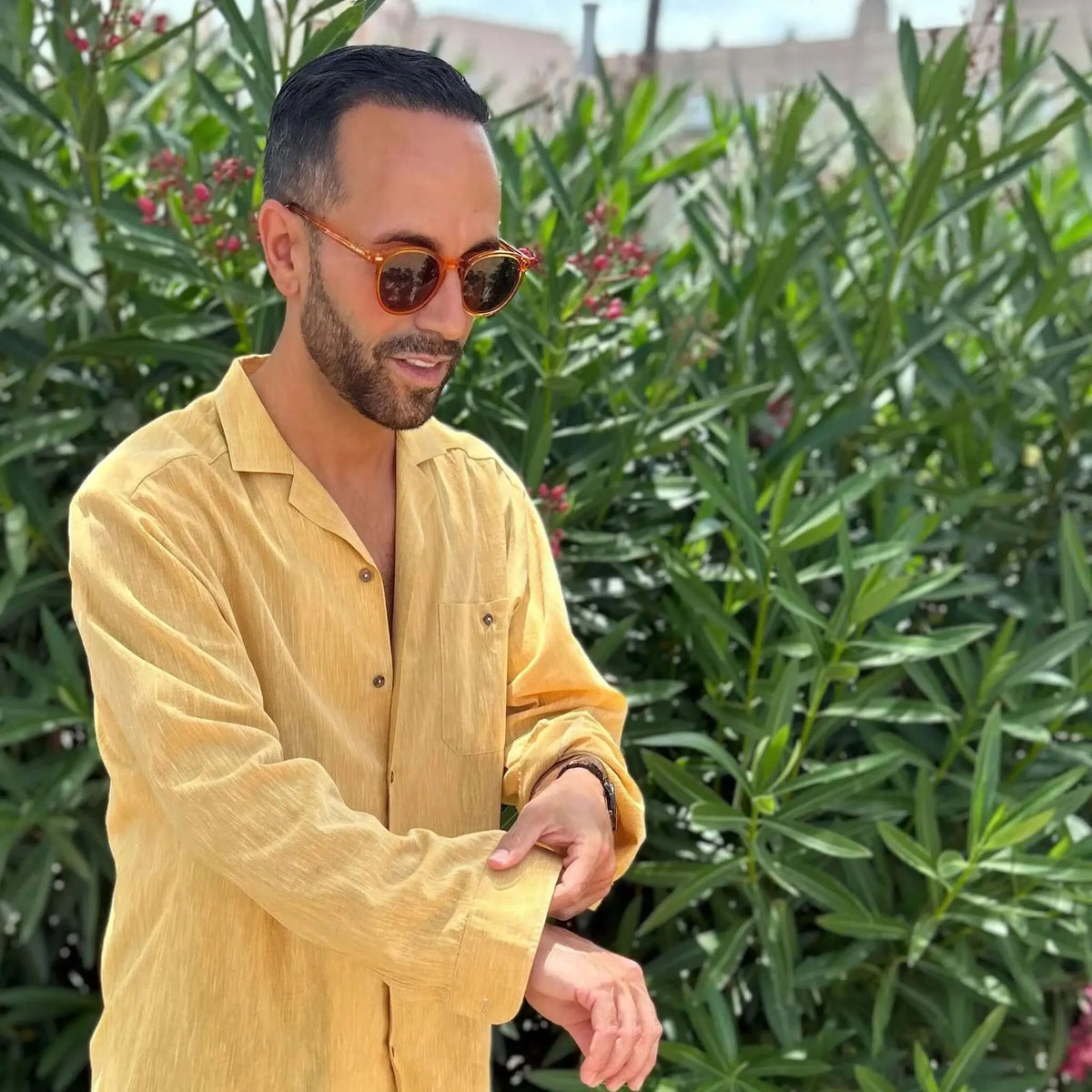 Hombre con camisa amarilla y gafas de sol de pasta naranja cristal, estilo moderno, posando frente a plantas verdes.