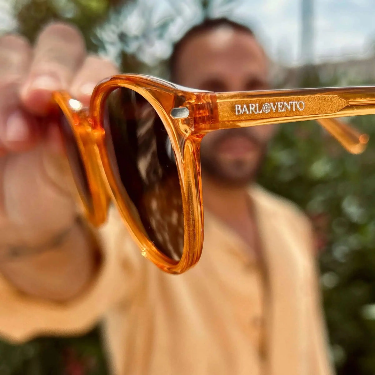 Gafas de sol de montura redonda en acetato naranja cristal brillante sostenidas por un hombre en exterior.