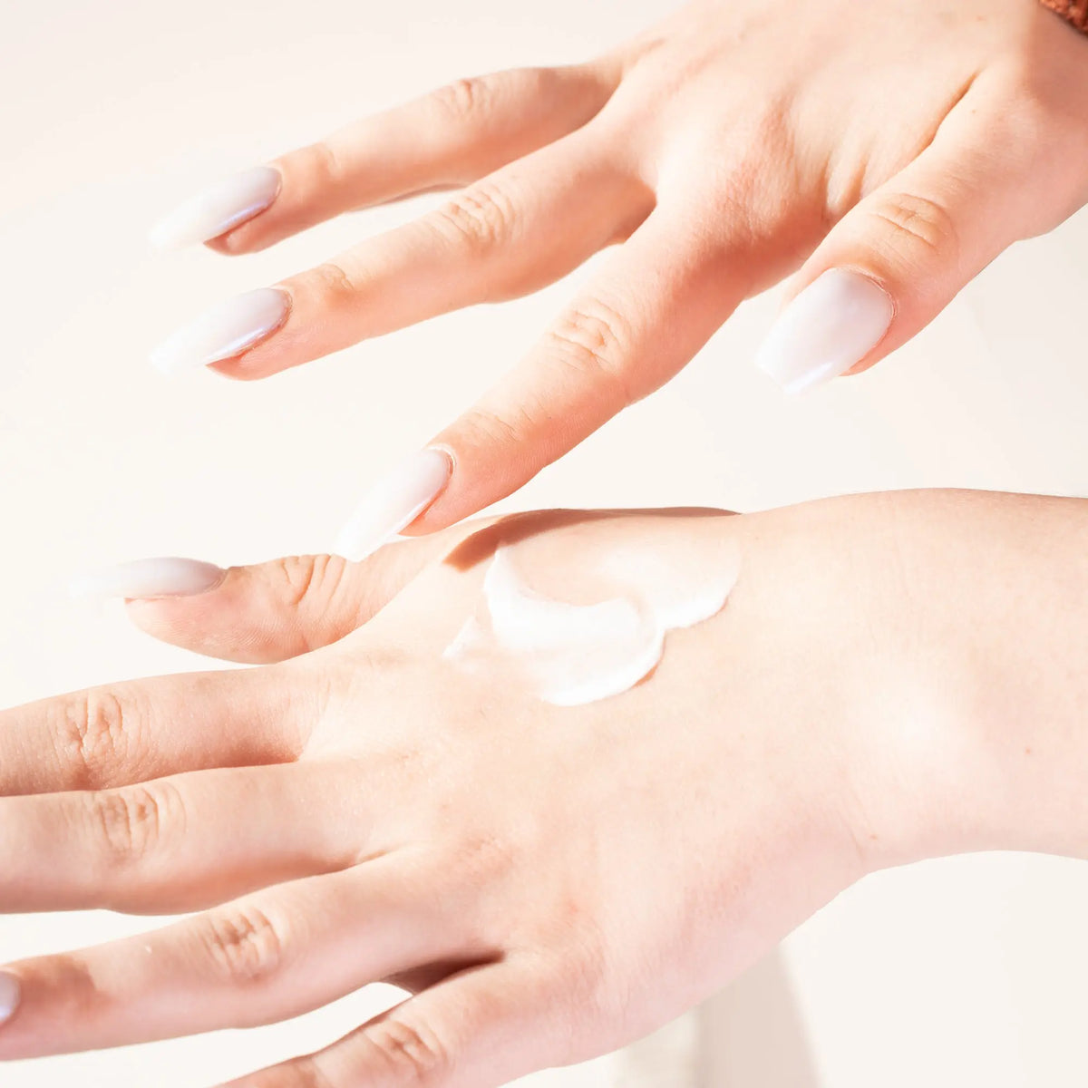 Creamy white hand cream dabbed on skin, hands with long glossy white nails against a soft neutral background.