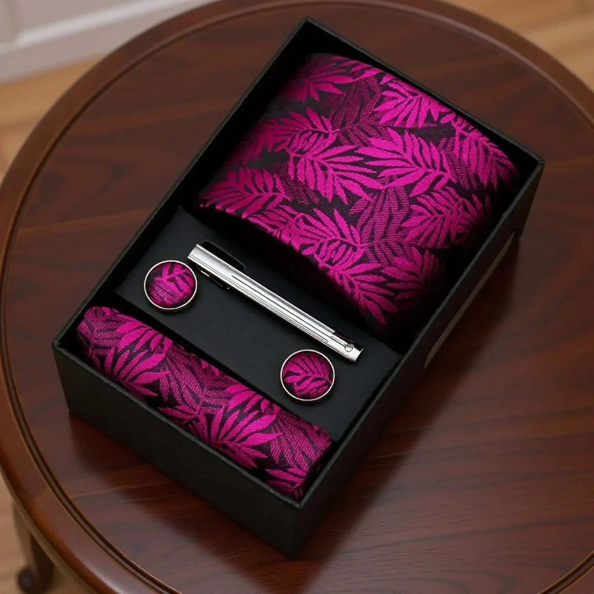 Set de corbata, pañuelo, gemelos y pasador magenta con estampado de hojas en caja sobre mesa de madera.