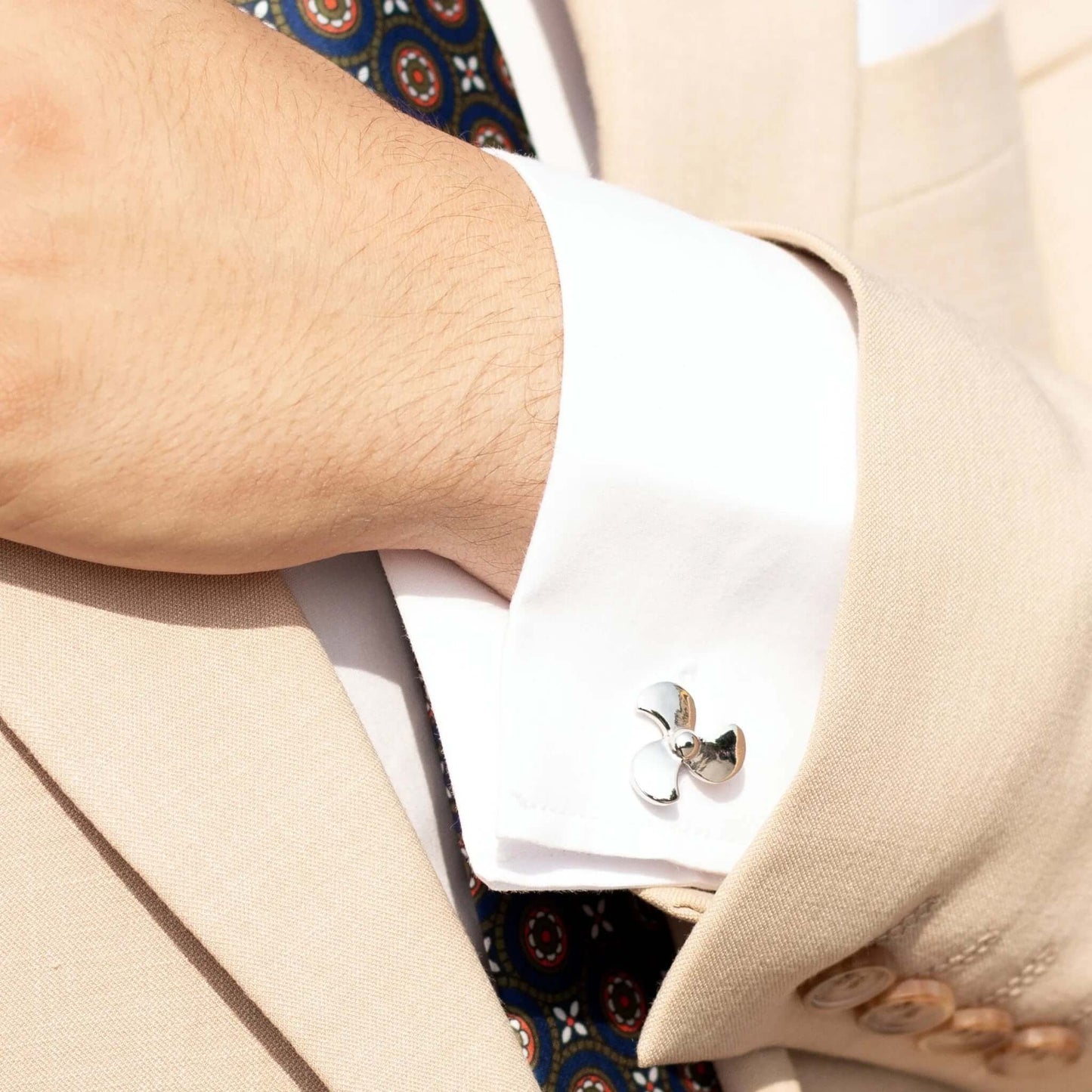 Gemelos de camisa plateados con forma de hélice de barco en traje beige, estilo elegante y detalle náutico.