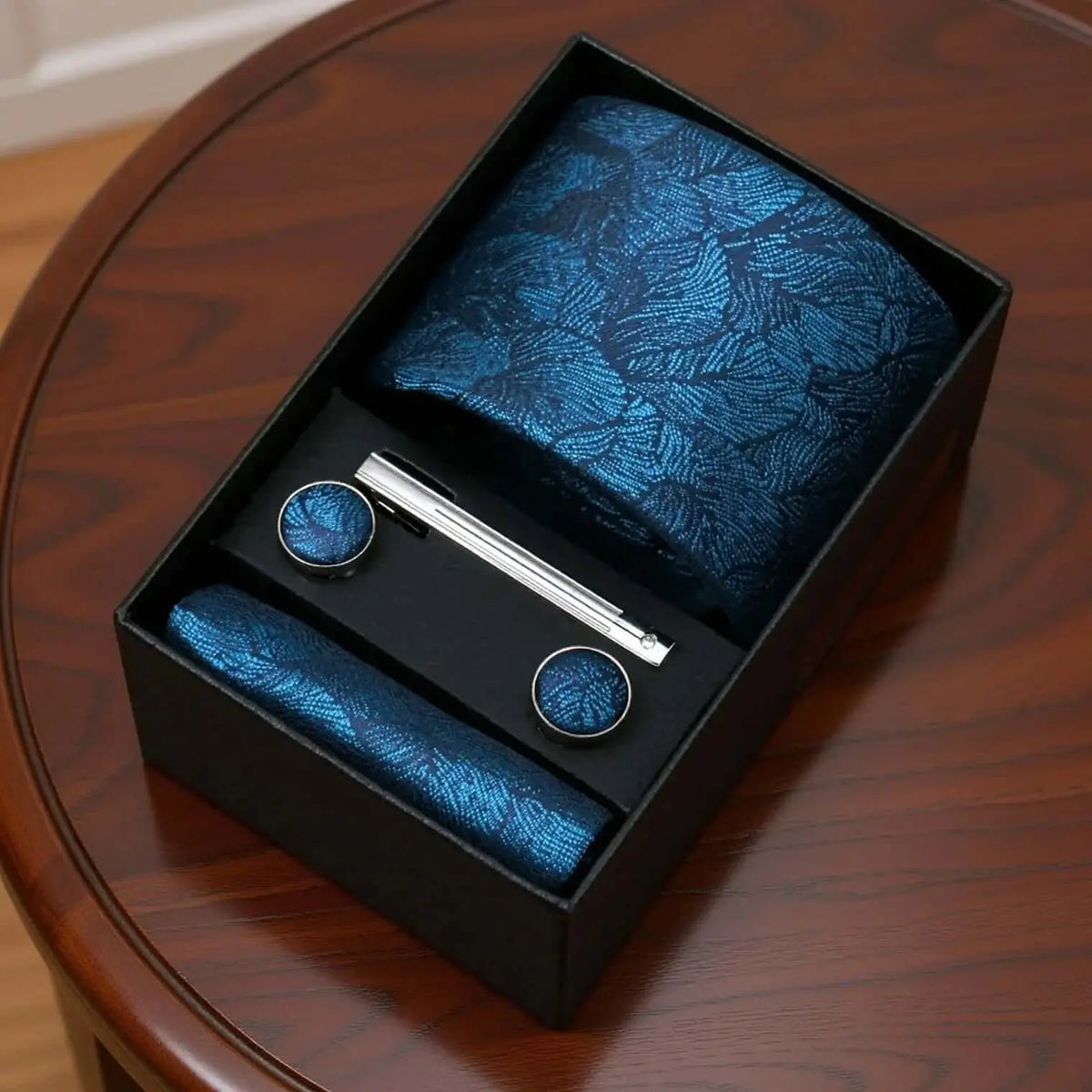 Set de regalo: corbata azul con patrón de hojas, pañuelo, gemelos y pasador metálico en caja negra sobre mesa de madera.