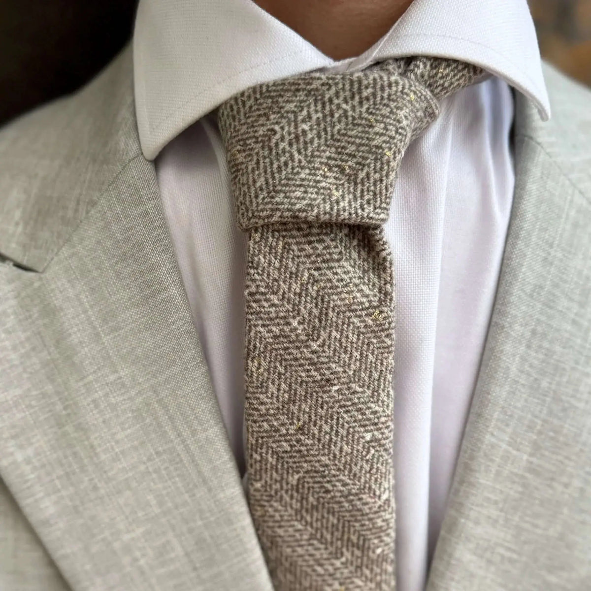 Corbata de lana beige con patrón de espiga y sutiles destellos dorados, anudada sobre camisa blanca y traje gris.