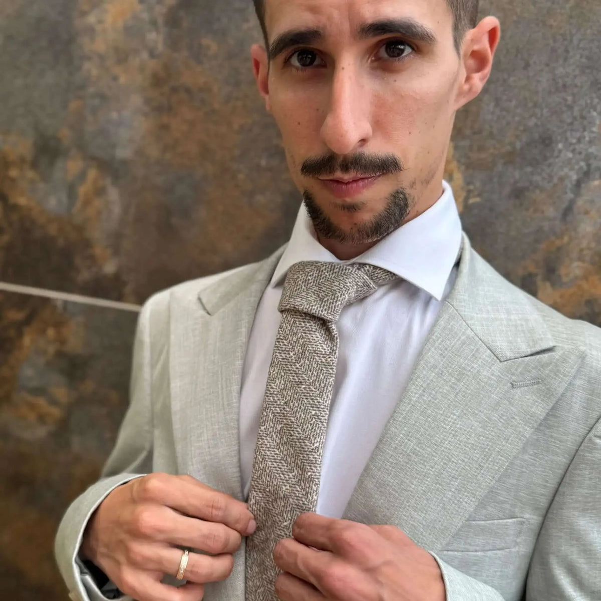 Corbata beige de lana con textura de espiga y sutil detalle dorado, anudada sobre camisa blanca y americana clara.