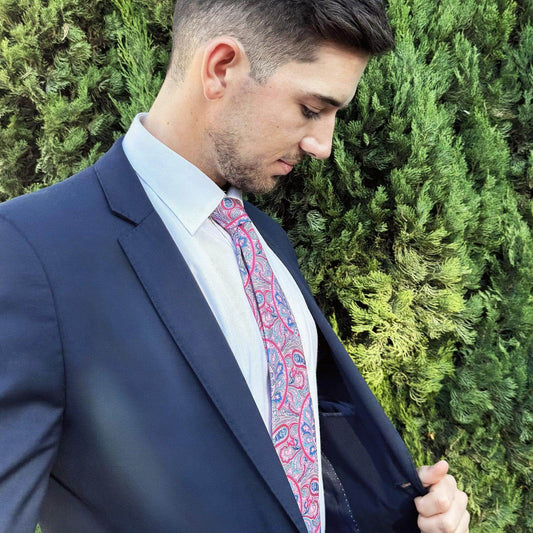 Hombre con traje azul, camisa blanca y corbata de seda rosa con estampado paisley sobre fondo de setos verdes.