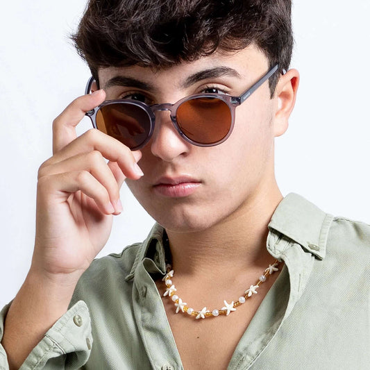 Joven con gafas de sol redondas de montura gris cristal y lentes marrones, usando camisa verde y collar de estrellas.
