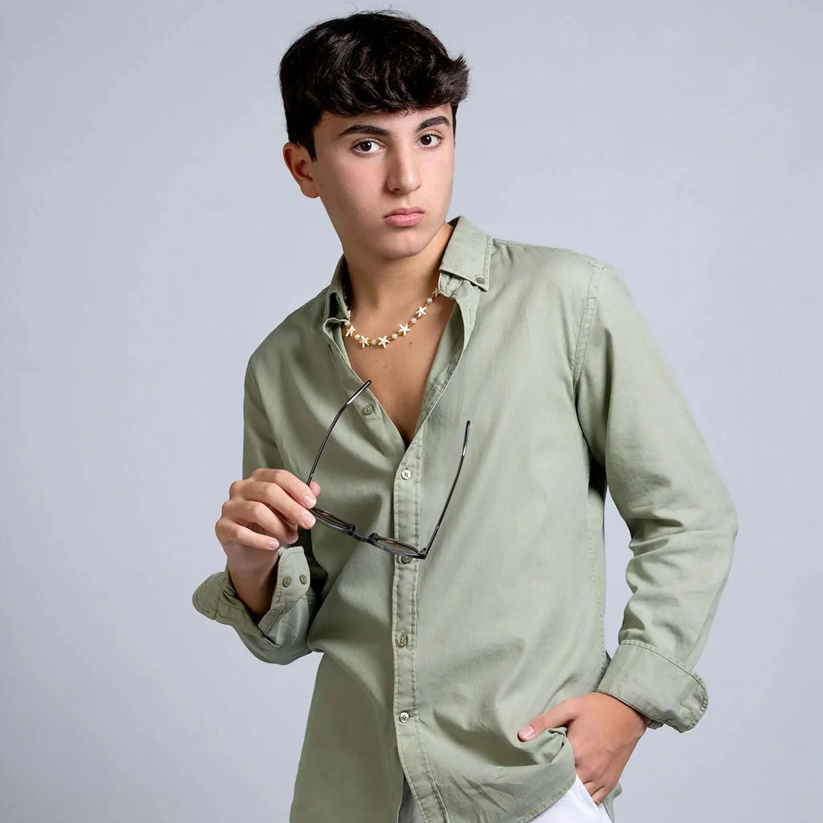 Joven con camisa verde sostiene gafas de sol redondas con montura de color gris cristal transparente.