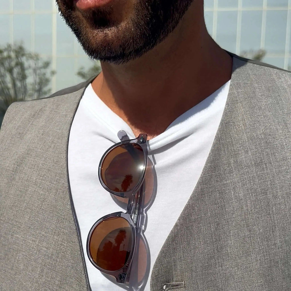 Gafas de sol redondas con montura gris cristal y cristales oscuros, colgadas en camiseta blanca de hombre con chaleco.