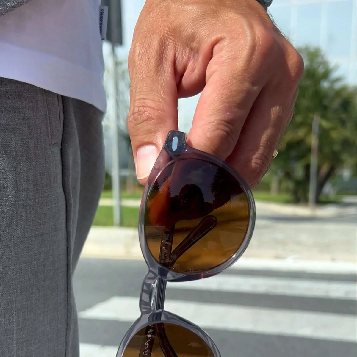 Gafas de sol redondas con montura gris cristal translúcida y lentes oscuras, sostenidas por hombre en entorno urbano.