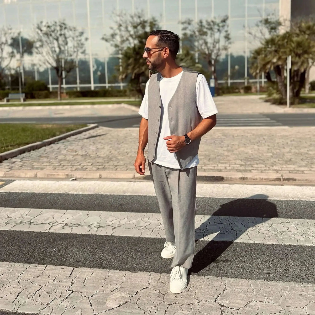 Hombre con traje gris y gafas de sol redondas de montura gris cristal caminando por un paso de cebra urbano.