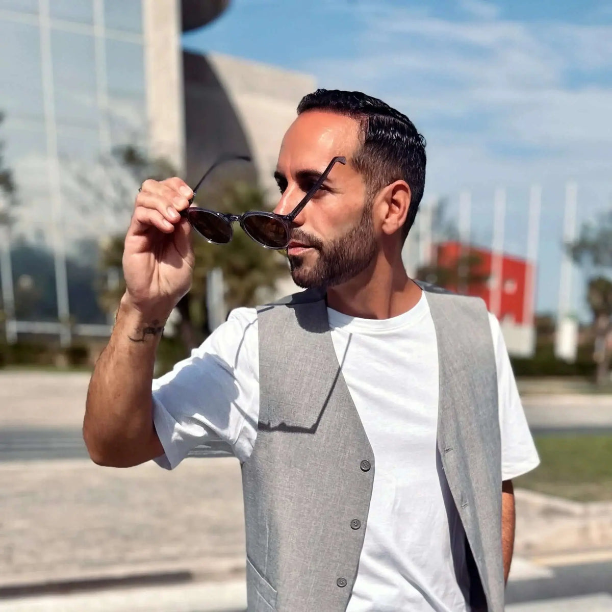 Hombre con chaleco gris sostiene gafas de sol redondas con montura de acetato gris cristal y lentes oscuras.