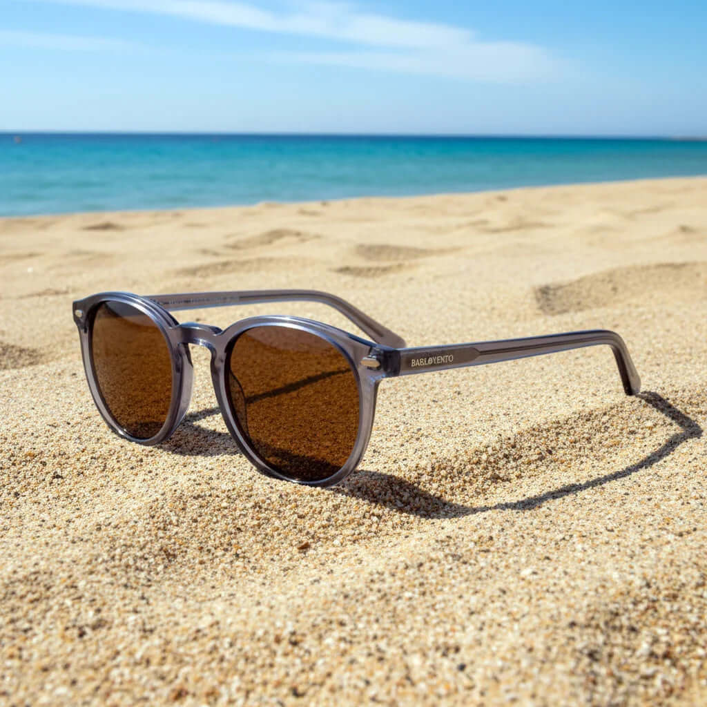 Gafas de sol redondas con montura gris cristal y cristales marrones sobre la arena de una playa soleada.