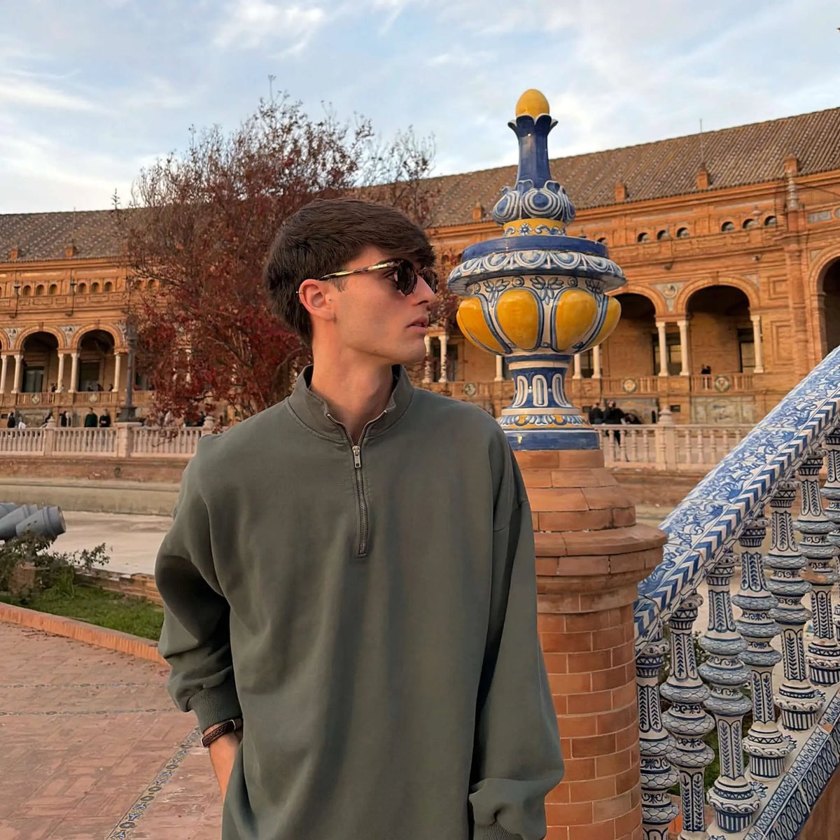 Joven con gafas de sol redondas de carey y sudadera verde, posando en plaza histórica. Estilo clásico y sofisticado.