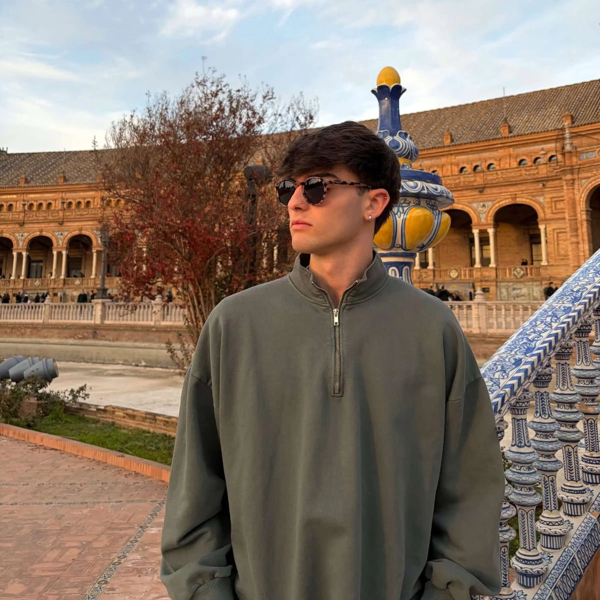 Joven con sudadera gris y gafas de sol redondas de pasta con estampado carey en un entorno arquitectónico histórico.