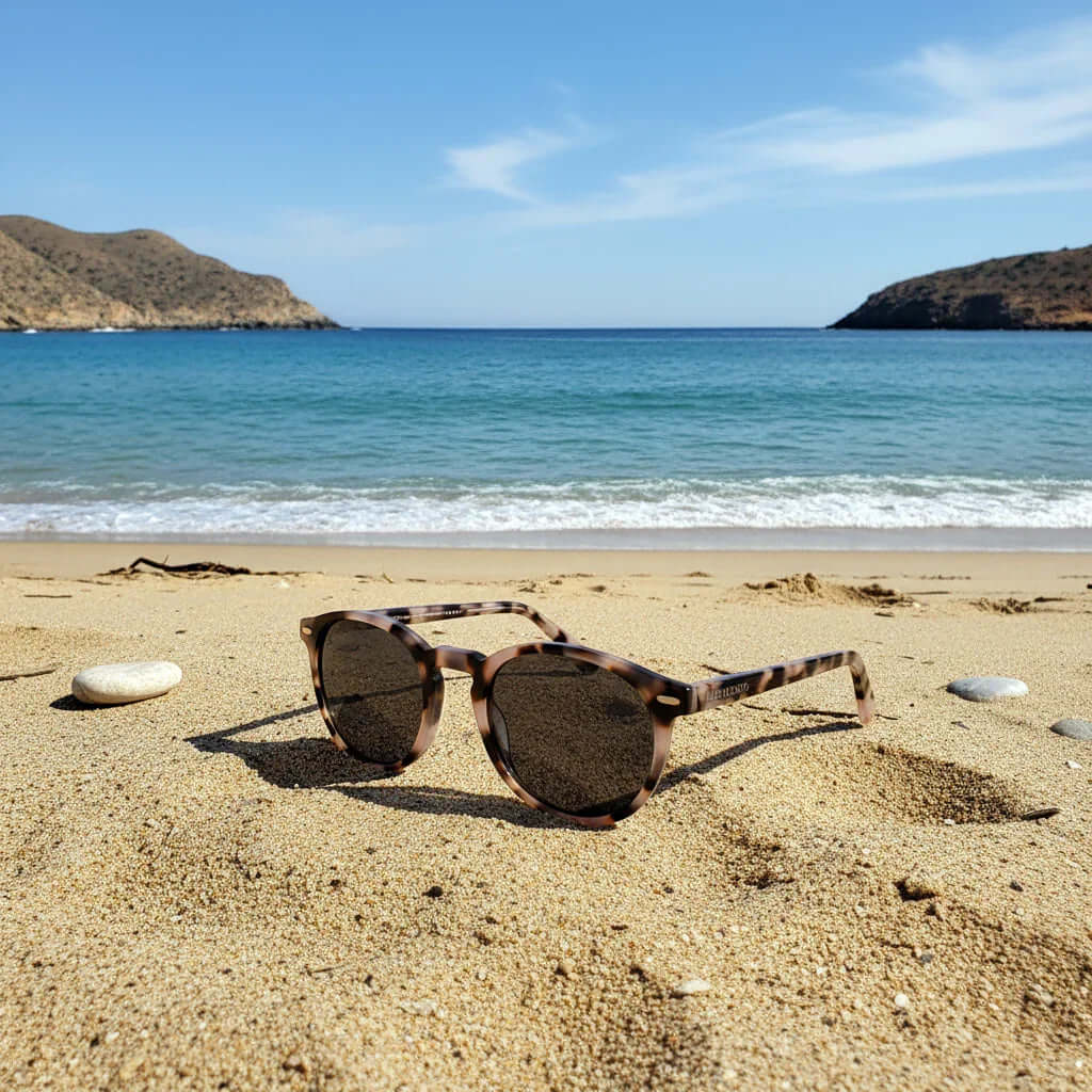 Gafas de sol redondas con montura de carey sobre la arena de la playa con el mar y montañas de fondo.