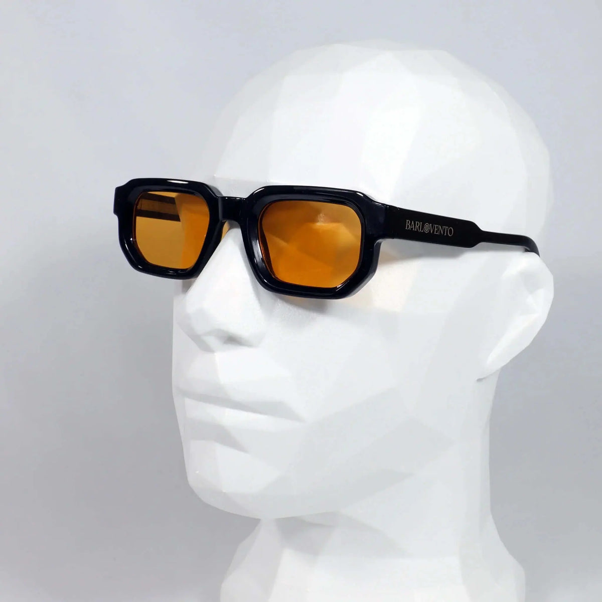Gafas de sol negras de estilo rectangular grueso con lentes naranjas, puestas sobre un maniquí blanco geométrico.