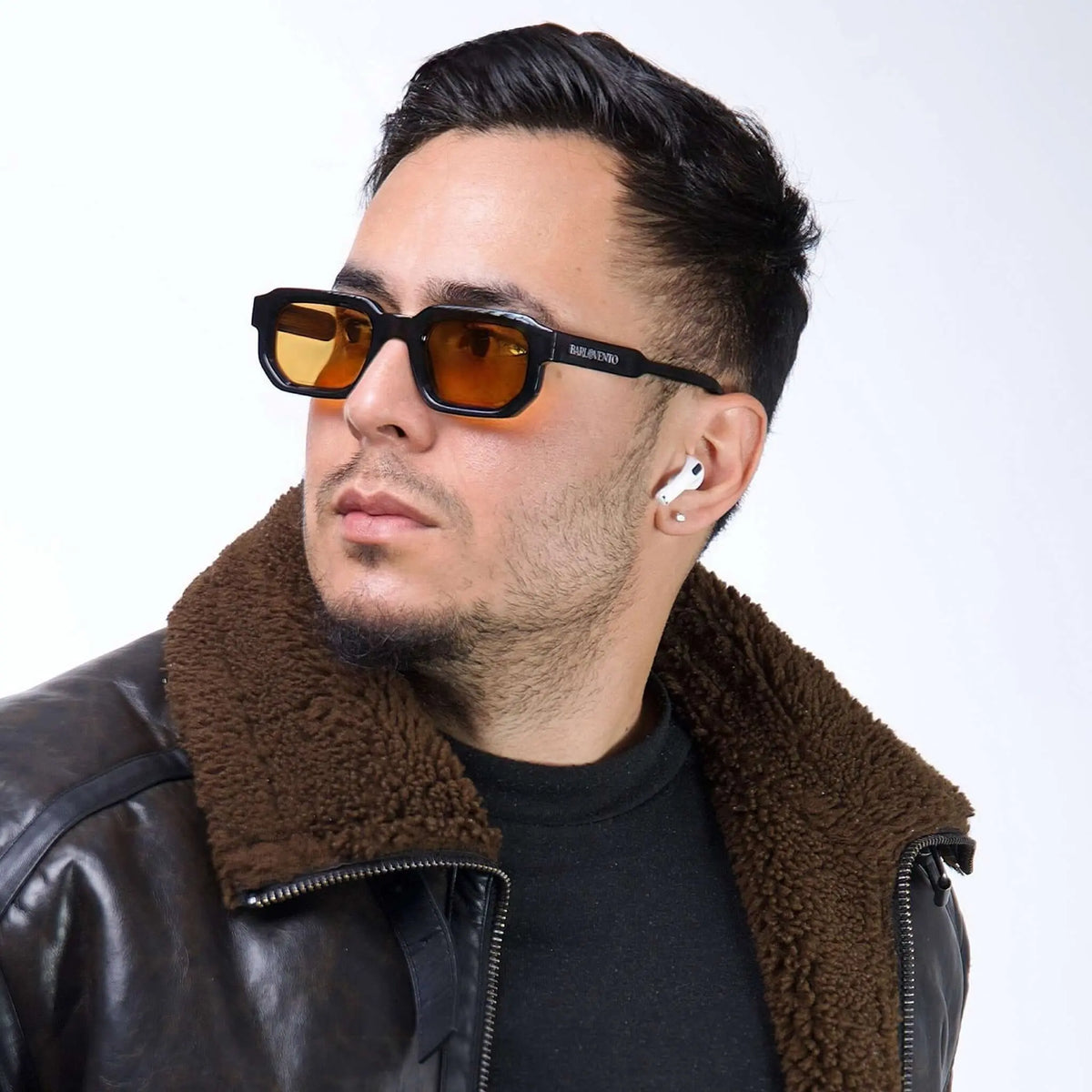 Hombre con gafas de sol negras de montura gruesa y lentes naranjas, usando chaqueta de cuero con cuello de borrego.