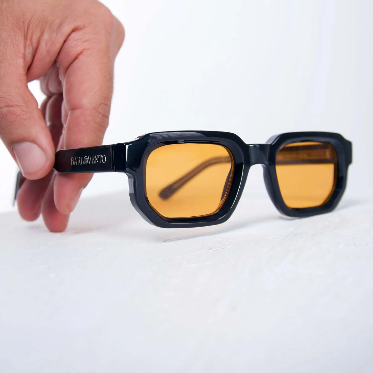 Gafas de sol rectangulares de montura negra brillante y cristales naranjas sostenidas por una mano sobre fondo blanco.