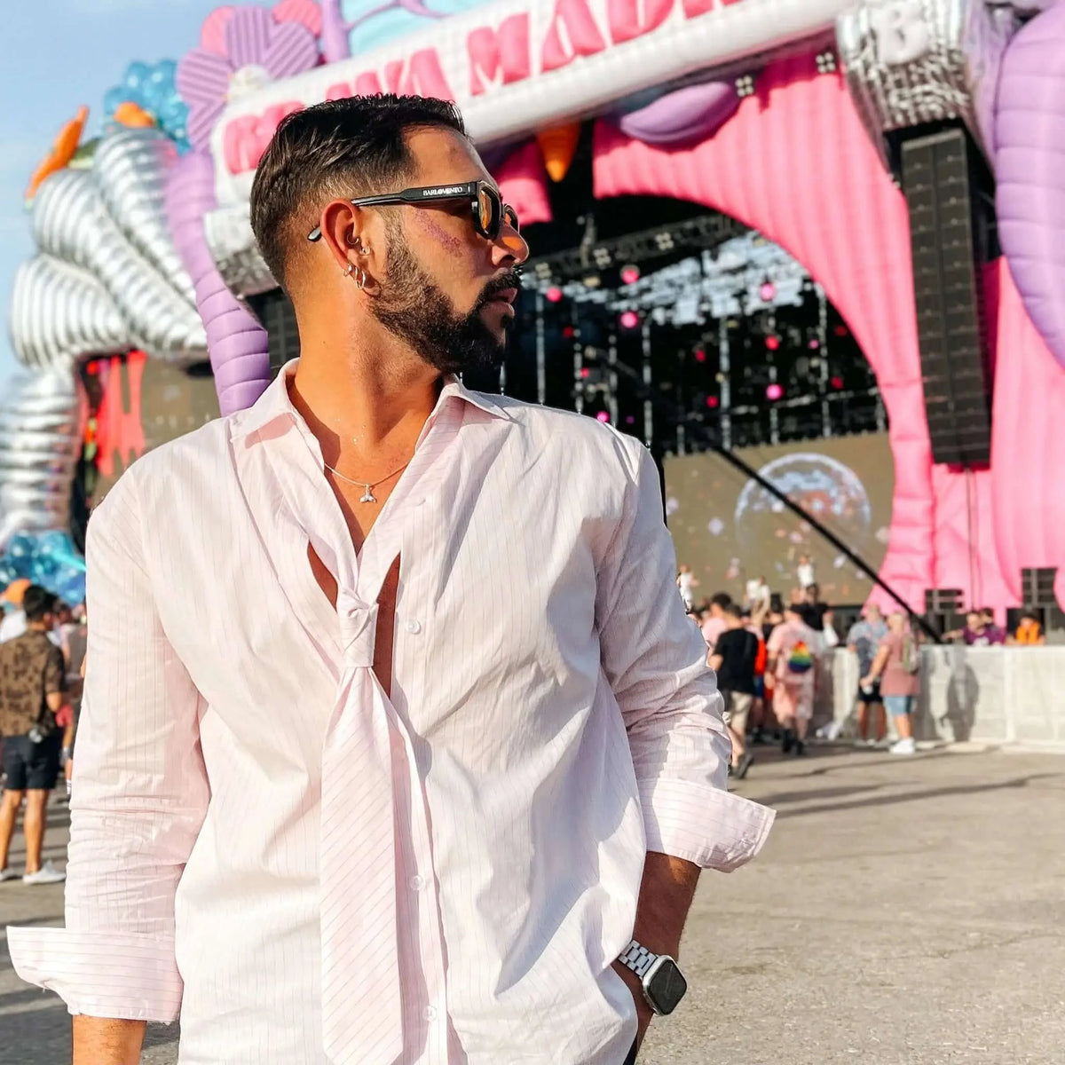 Hombre con camisa rosa y gafas de sol negras de montura gruesa con lentes naranjas en un festival al aire libre.