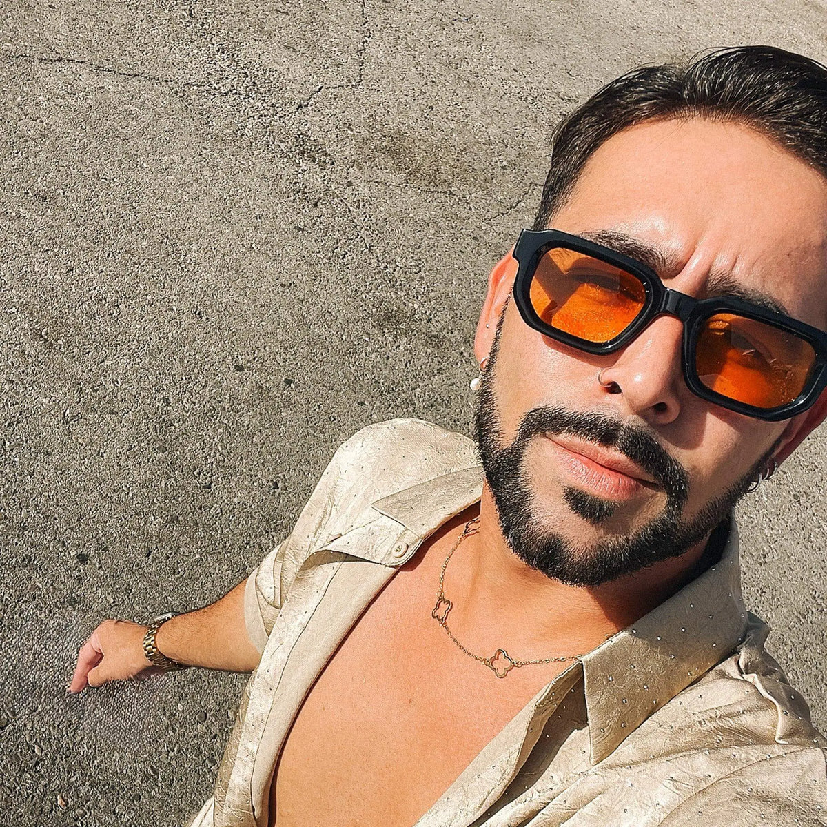 Hombre con gafas de sol cuadradas de montura negra y lentes naranjas, vistiendo camisa satinada color champagne.