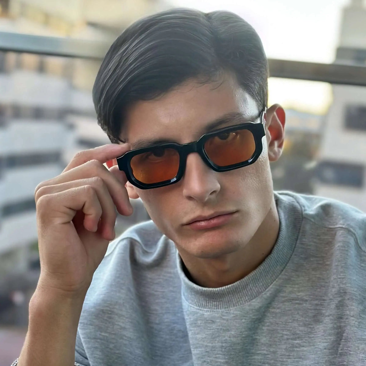 Joven con sudadera gris usa gafas de sol de montura negra rectangular y lentes naranjas estilo moderno y urbano.