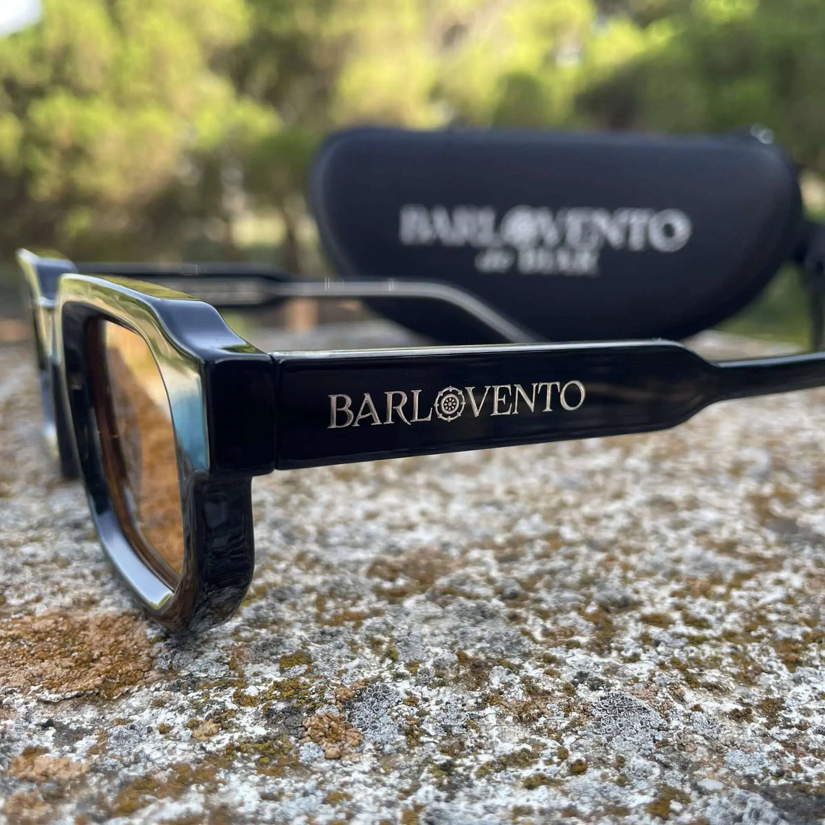 Gafas de sol negras de estilo grueso con lentes naranjas y acabado brillante sobre superficie de piedra natural.