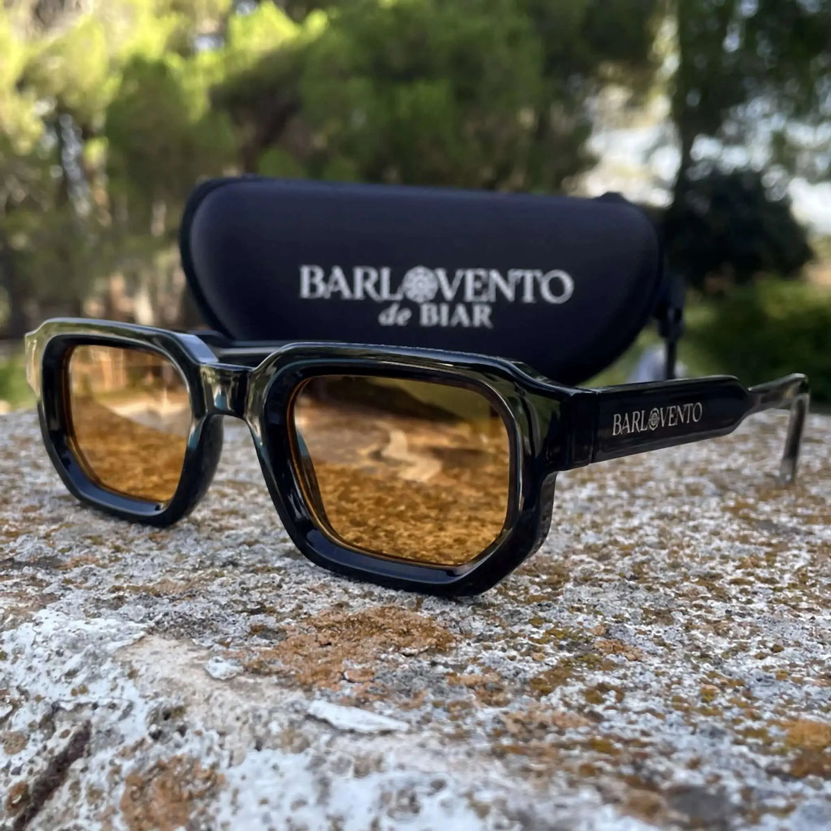Gafas de sol negras de estilo rectangular con montura gruesa y lentes naranjas, sobre una superficie de piedra rugosa.