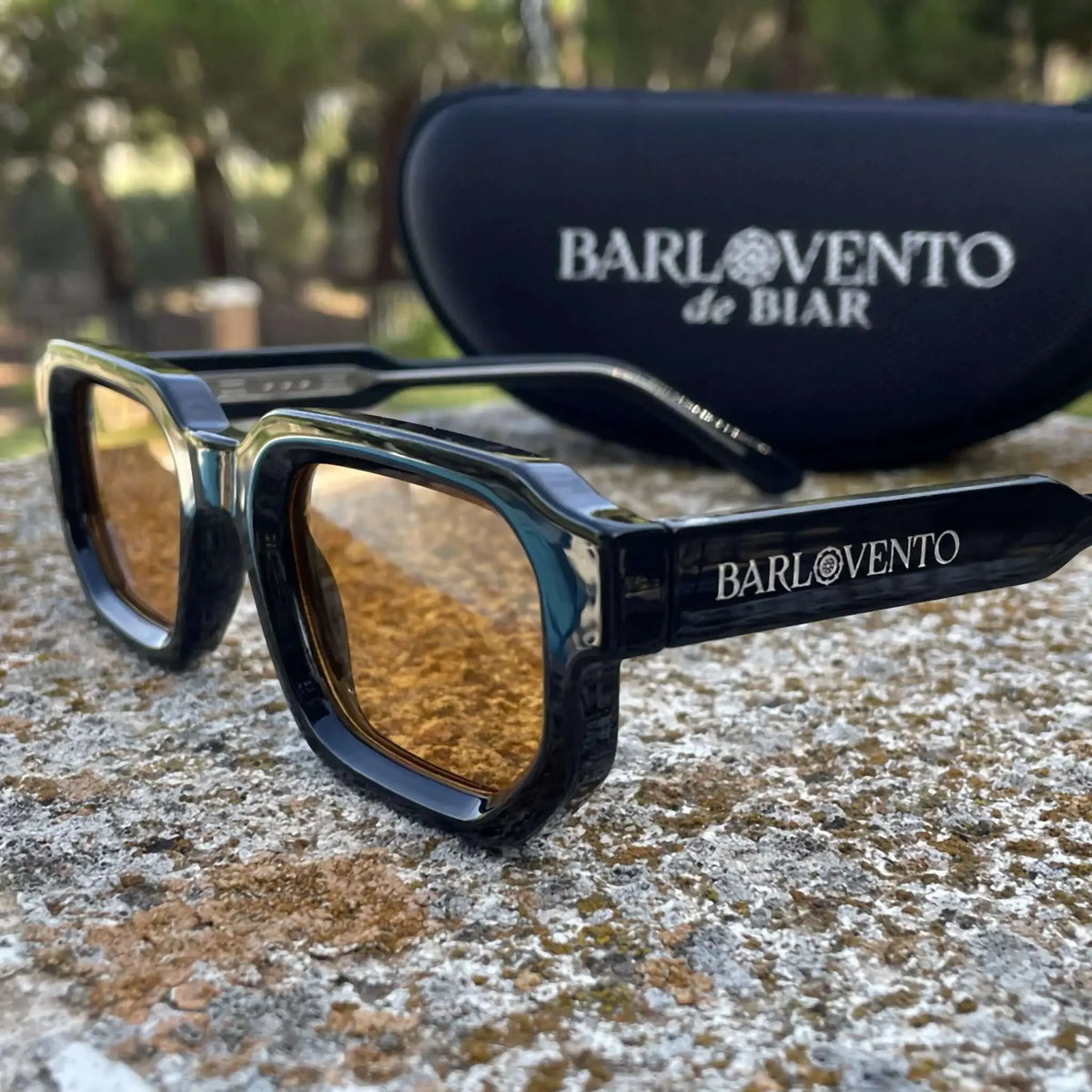 Gafas de sol negras de estilo rectangular con montura gruesa y lentes color naranja sobre una superficie de piedra.