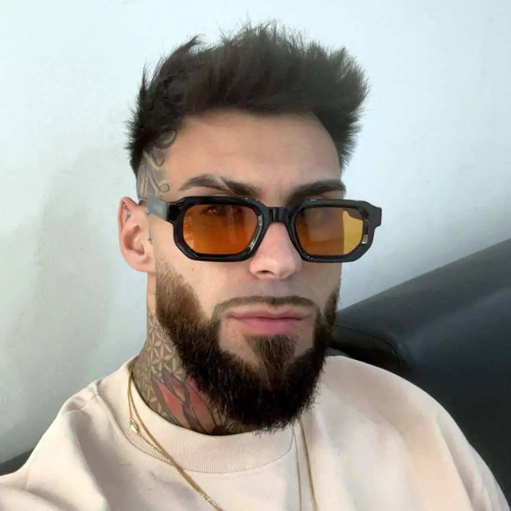 Hombre con gafas de sol rectangulares negras y lentes naranjas, estilo moderno y audaz, con barba y tatuajes en cuello.