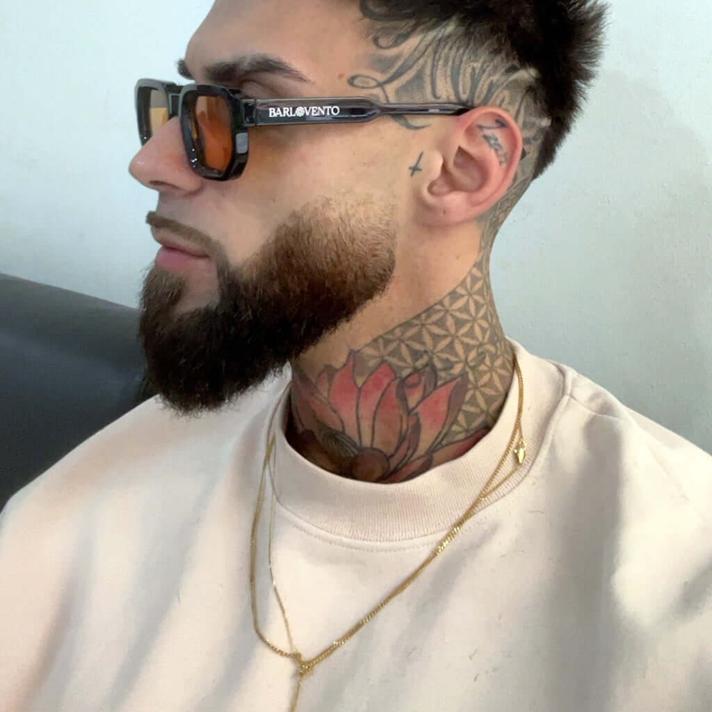 Hombre tatuado con gafas de sol negras de montura cuadrada gruesa y lentes naranjas en estilo urbano moderno.