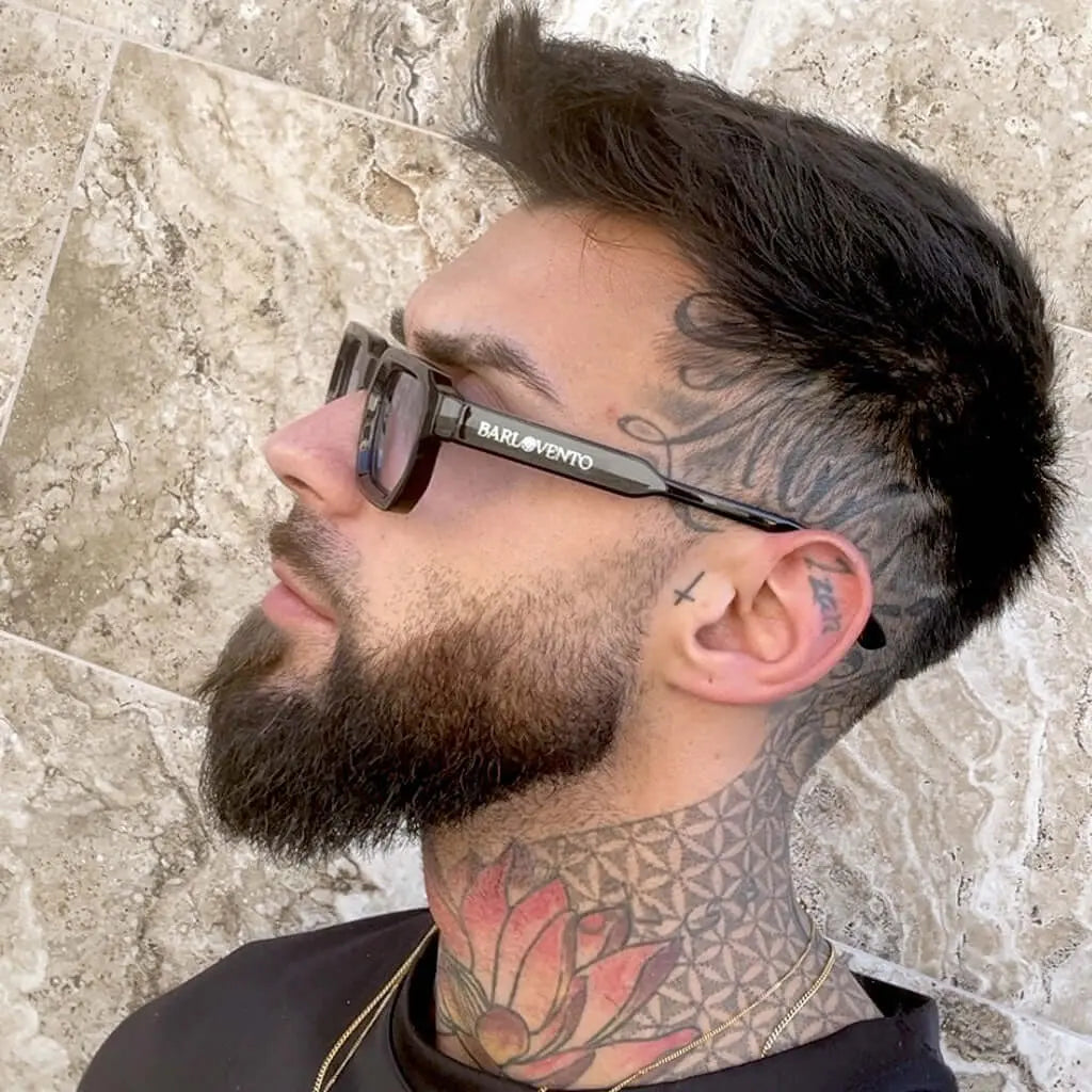 Hombre tatuado con barba usando gafas de sol negras de montura cuadrada gruesa y lentes naranjas con estilo moderno.