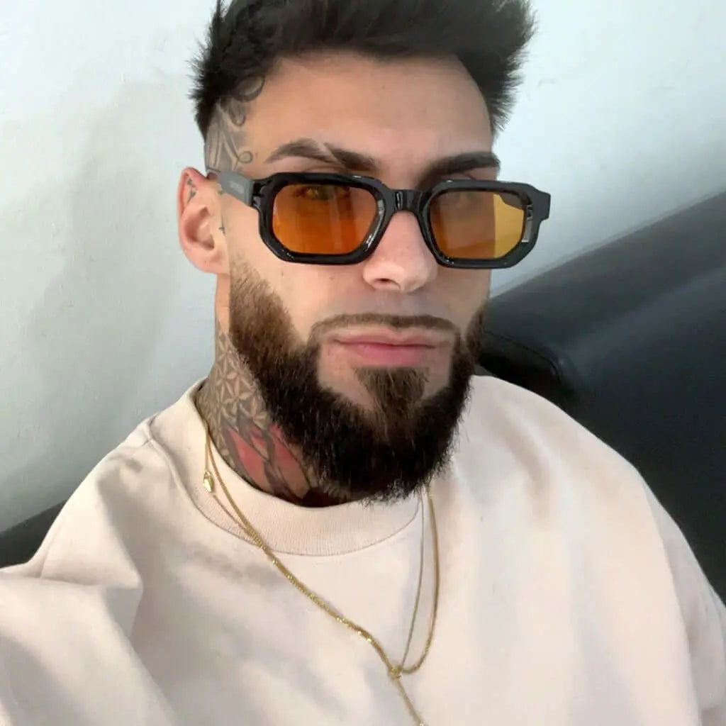 Hombre con barba y gafas de sol rectangulares de montura negra y lentes naranjas, estilo moderno y urbano.