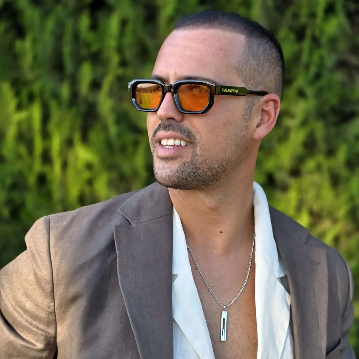 Hombre con gafas de sol cuadradas de montura negra brillante y lentes naranjas, vistiendo americana gris y camisa.