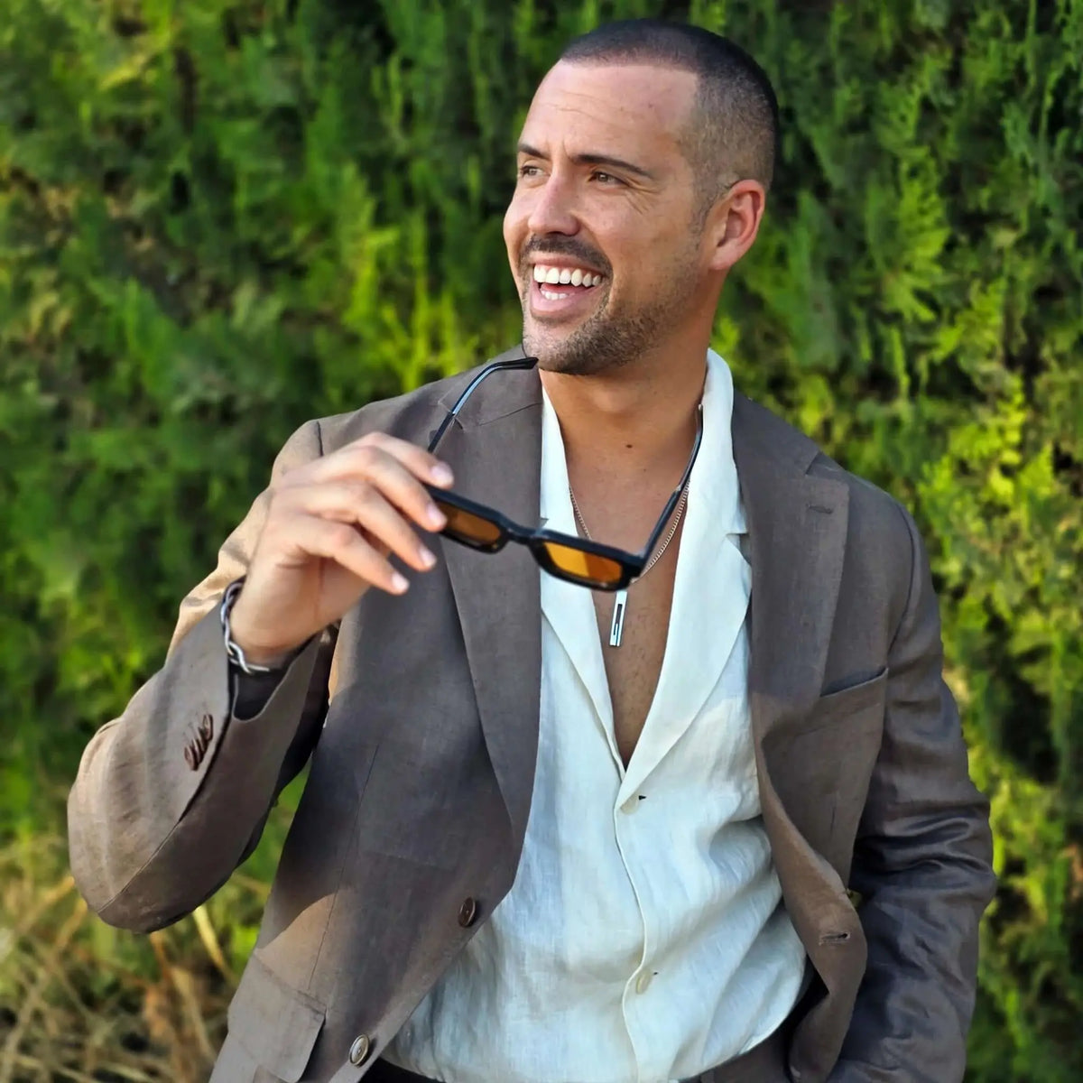 Hombre con americana marrón sostiene gafas de sol negras de estilo rectangular con cristales naranjas.