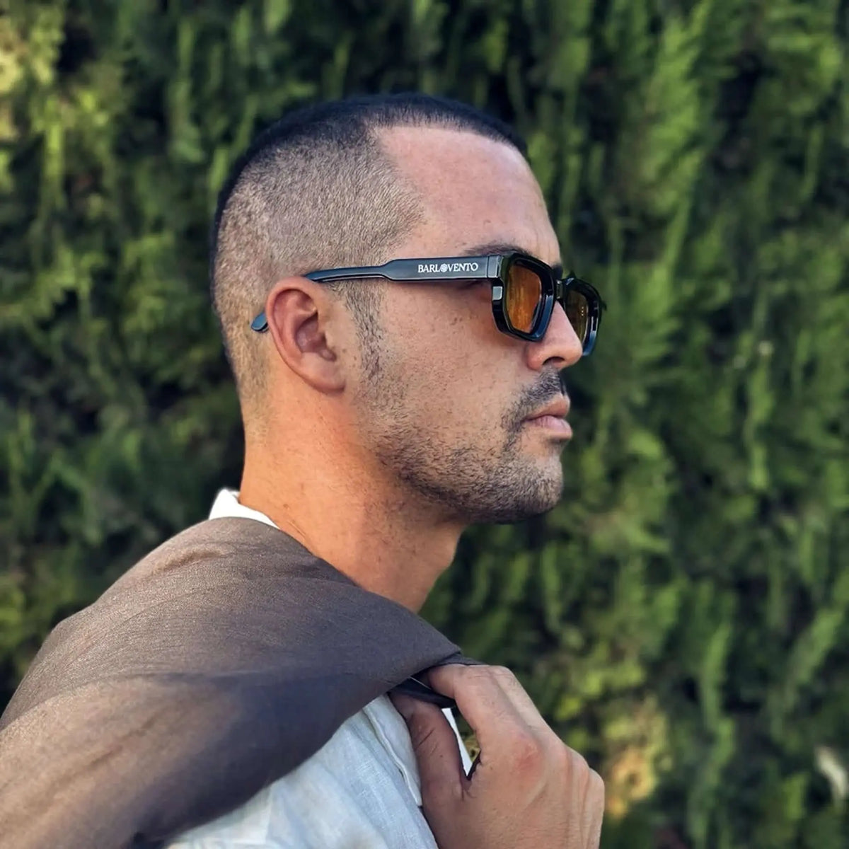 Hombre con gafas de sol negras de montura cuadrada y lentes naranjas, estilo moderno con fondo de follaje verde.
