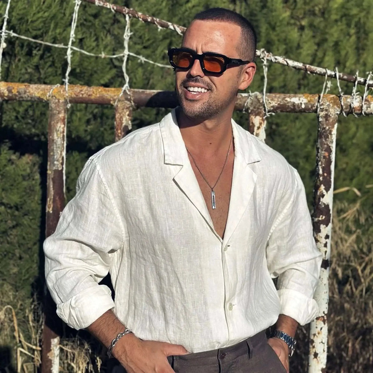 Hombre con camisa de lino y gafas de sol negras de montura gruesa y lentes naranjas en un entorno exterior.