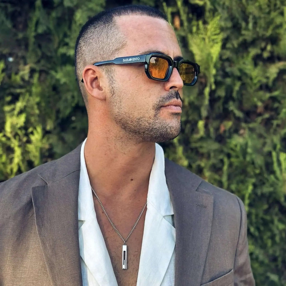 Hombre con gafas de sol negras de montura cuadrada y lentes naranjas, traje marrón y camisa blanca al aire libre.