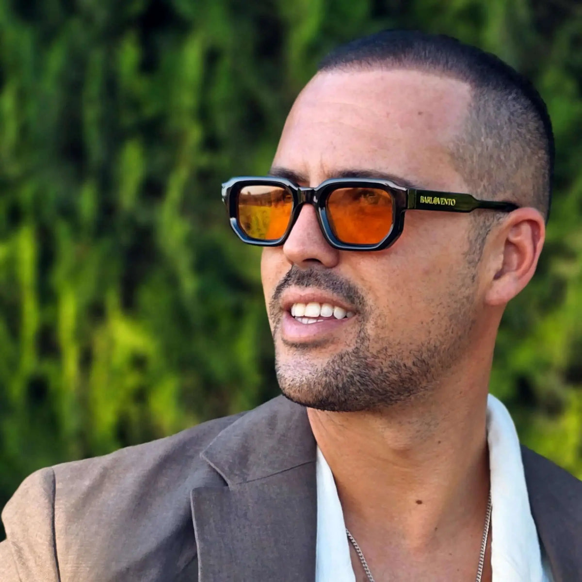 Hombre con blazer gris y gafas de sol de montura negra cuadrada y lentes naranjas llamativas en entorno natural.